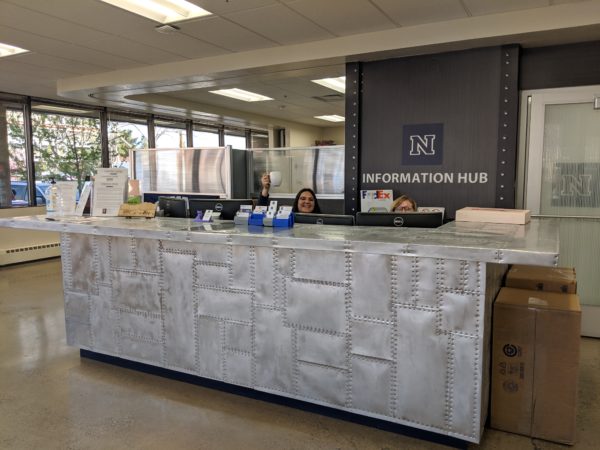 Two girls just visible over the top of a tall metal desk with a sign behind them that says "Information Hub" with the University of Nevada, Reno N above it. It is a reception desk.