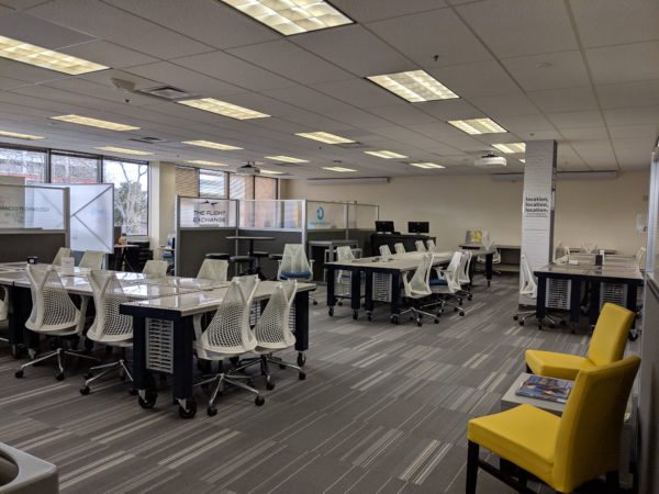 A large coworking space with numerous metal tables, white chairs, and grey cubicle dividers. On the right are two small yellow chairs with a small coffee table between the two.